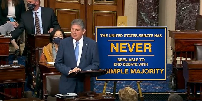 Manchin stands next to a sign
that says 'The United States Senate has never been able to end debate with a simple majority.'