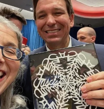 DeSantis holds a cut-out snowflake
and stands next to a voter while wearing an unpersuasive smile