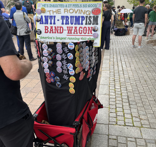 A guy is selling pins out of a wagon labeled 'The Roving Anti-Trumpism Bandwagon,' which also has a banner that says 'He's indicted and it feels so good'