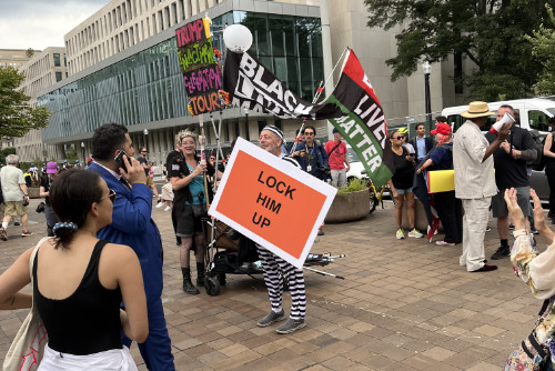 A guy in an old-style striped prison suit holds a sign that says 'Lock him up' while flanked by Black Lives Matter flags