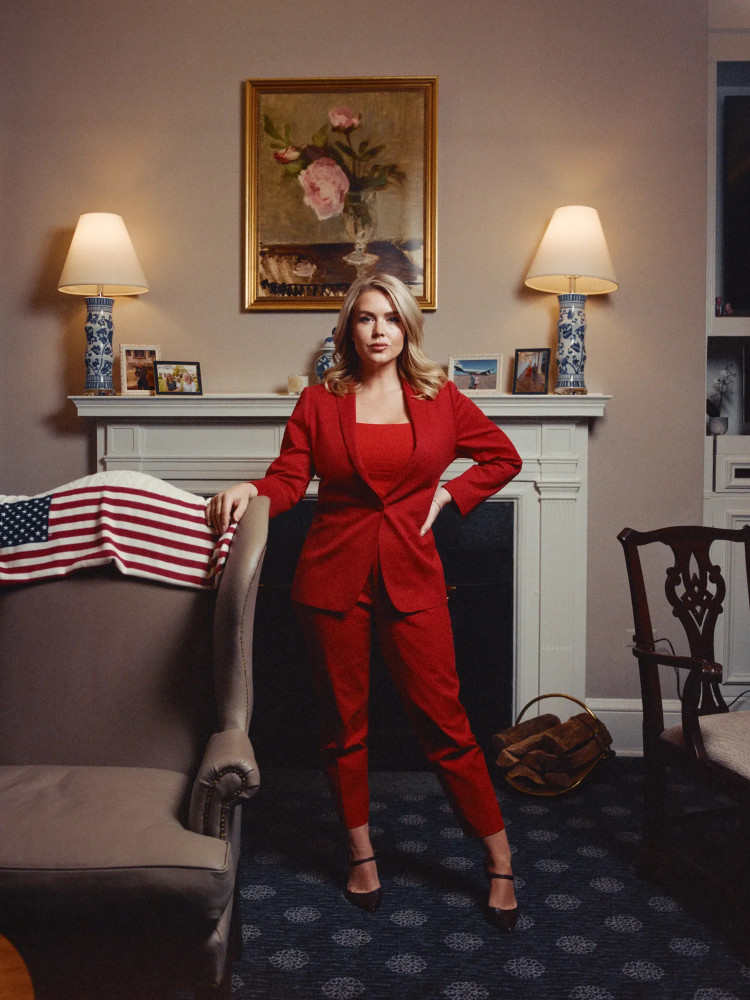 Karoline Leavitt stands in front of a fireplace, next to am empty chair