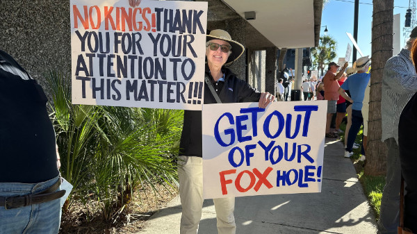 A woman, amid a sizable
crowd holds signs that say 'NO KINGS! THANK YOU FOR YOUR ATTENTION TO THIS MATTER!' and 'GET OUT OF YOUR FOX HOLE!' with
FOX in red letters and the rest in blue.