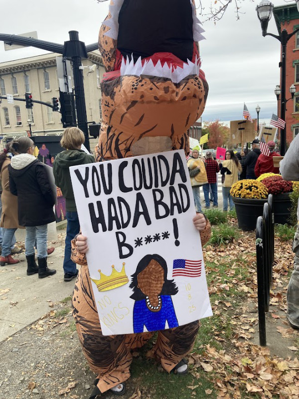 A person in a
dinosaur costume holds a sign with a drawing of Kamala Harris that says 'YOU COULDA HAD A BAD B****'