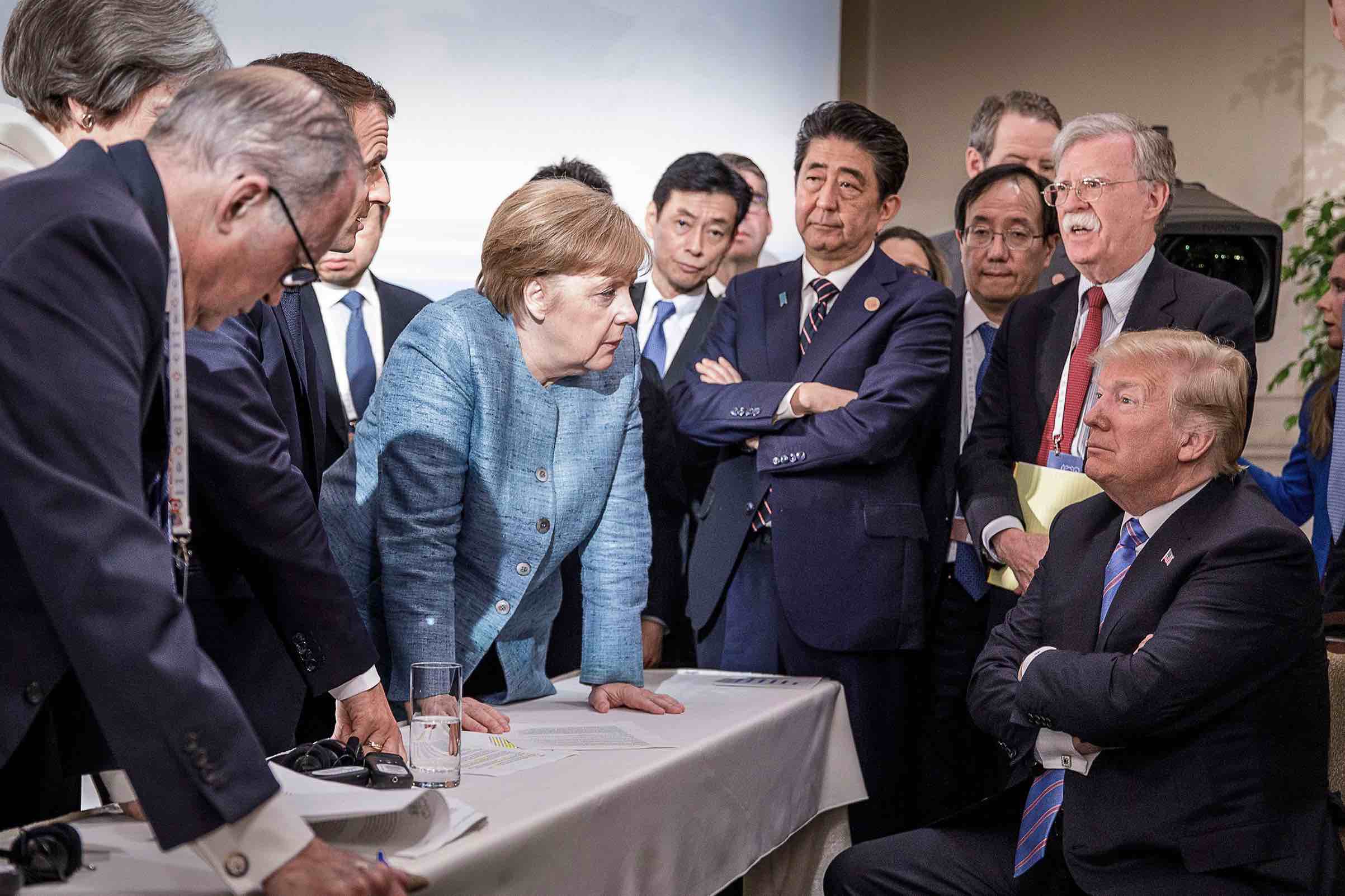 Angela Merkel stands and looks daggers at Trump, while Shinzo Abe is disdainful, with arms crossed, and Donald Trump is petulant, also with arms crossed