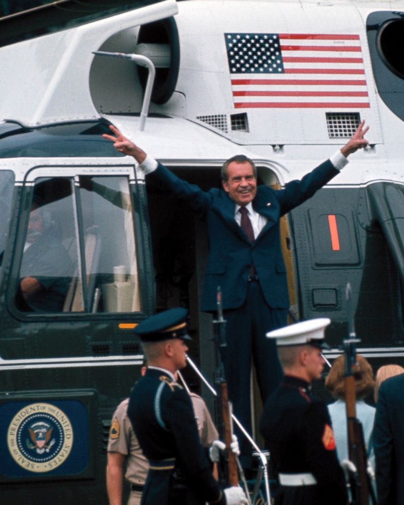 Nixon smiles and makes a double
victory sign, with both hands, while standing on the steps of a Marine Corps helicopter