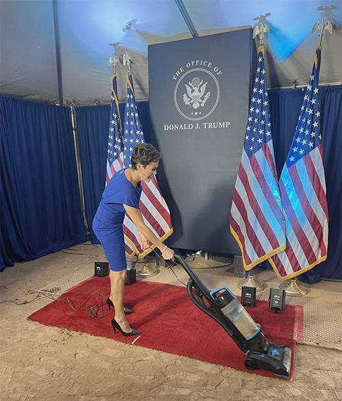 Lake vacuuming the carpet in front of a sign
that says 'Office of Donald Trump'