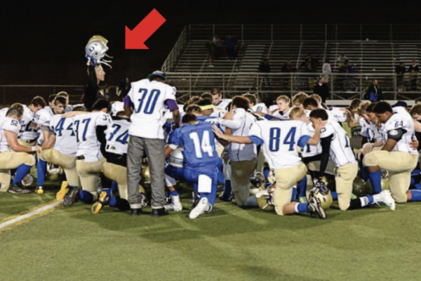 The coach stands among at least 60
people, including kneeling players from both teams, as indicated by their different-colored jerseys