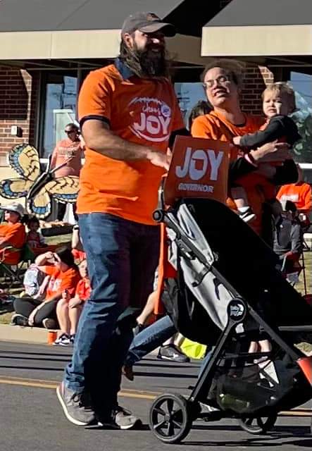 Two people wearing 
orange shirts and carrying an orange sign that say 'Joy'