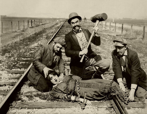 Three mustachioed men tie a
damsel in distress to train tracks
