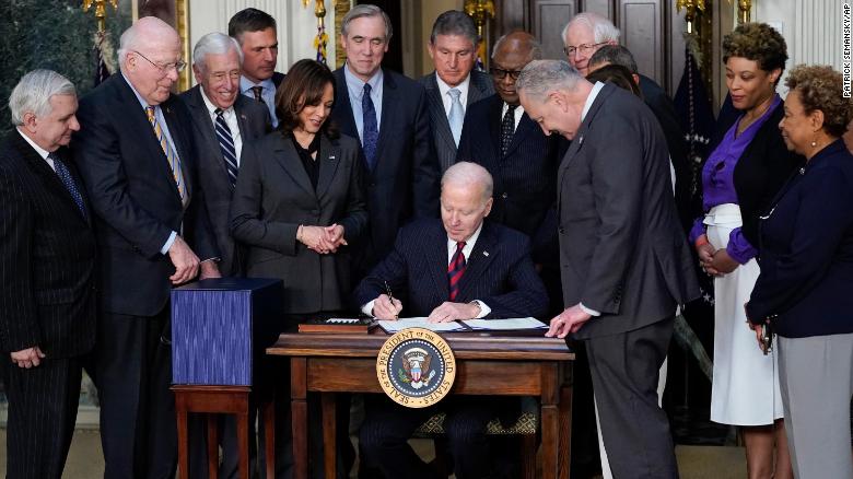 At least 14 politicians gather around the 
desk as Biden signs the law; not all are clearly visible, but Pat Leahy, Kamala Harris, Joe Manchin, Jim Clyburn, and Chuck Schumer
are all clearly shown; Schumer is slumped in a pose that makes it look like he's fallen asleep standing up.