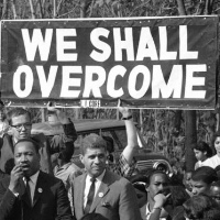 A sign, held by someone standing behind MLK Jr., says 'We Shall Overcome
