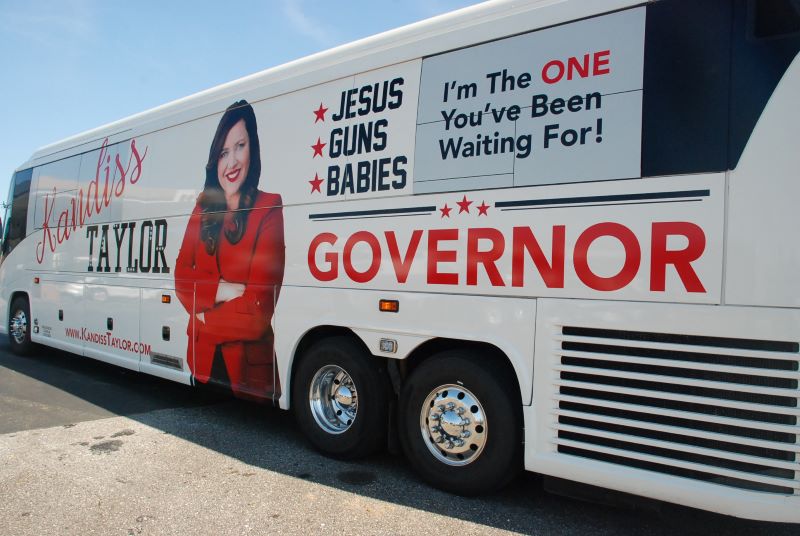 The bus reads 'Jesus. Guns. Babies.'
and also 'I'm the ONE you've been waiting for,' with 'ONE' in big, red capital letters.