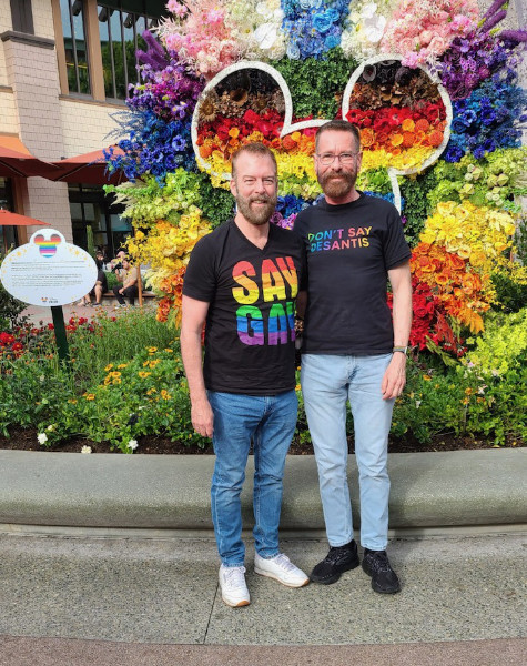 Two gay, bearded gentleman
stand in front of a Mickey Mouse made up of a rainbow of colored flowers
