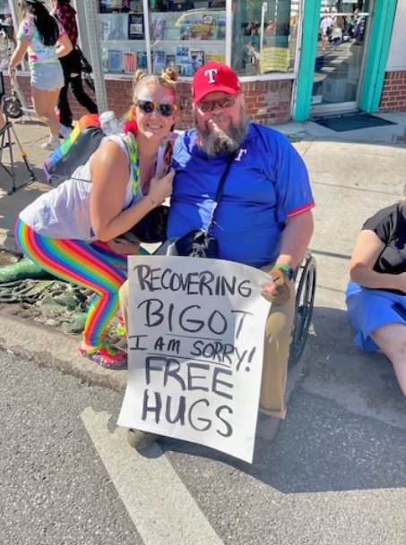 Nash sits in a wheelchair, holding 
a sign that says 'Recovering Bigot! I am Sorry! Free Hugs!' as a woman in rainbow pants takes him up on the offer.
