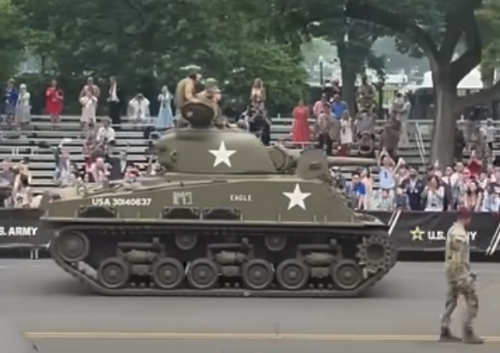 A soldier and
a tank are traversing the parade route; the grandstands they are walking past are less than half full