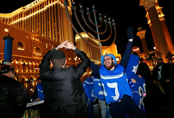 A man in a dreidel costume stands in front of a menorah, in front of a glitzy hotel done in Italianate style