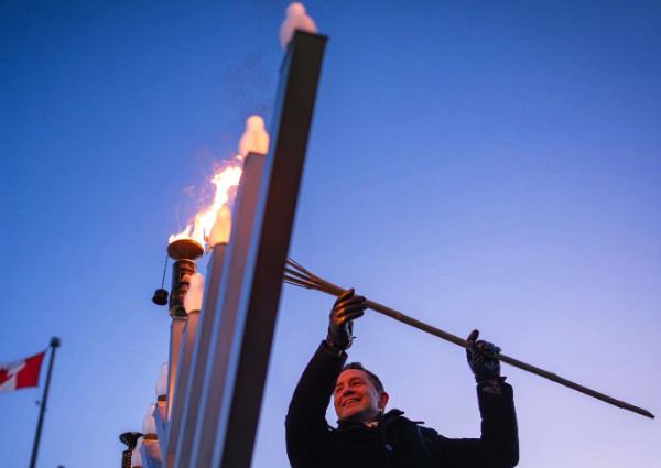 A man lights a giant menorah, a red and white flag with some sort of design on it is visible in the background