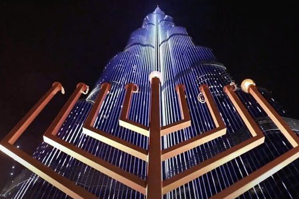 A view of a menorah looking up at it; behind is a VERY tall, tapered skyscraper in modernist style