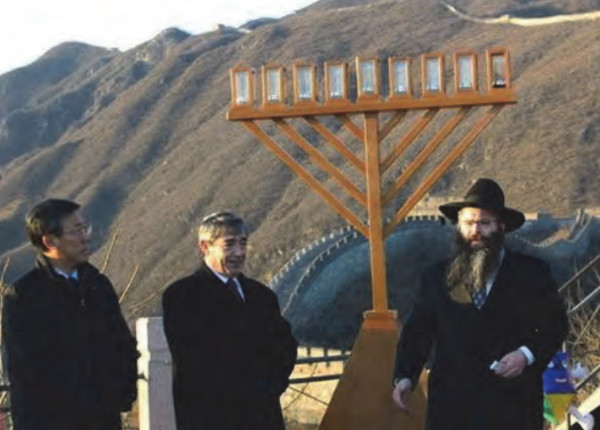 Two Asian men and a Jewish man light a menorah, there is a very, very long wall behind them