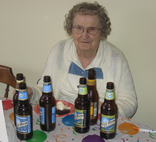 A smiling woman in her 80s, with numerous
bottles of beer on the table