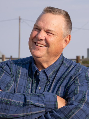 Jon Tester wearing a plaid shirt, standing in a field, and smiling