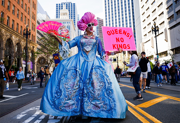 A person with a sign that says 'NO KINGS ONLY QUEENS'
and who is wearing a stylized 'queen' costume that suggests 'Alice in Wonderland'