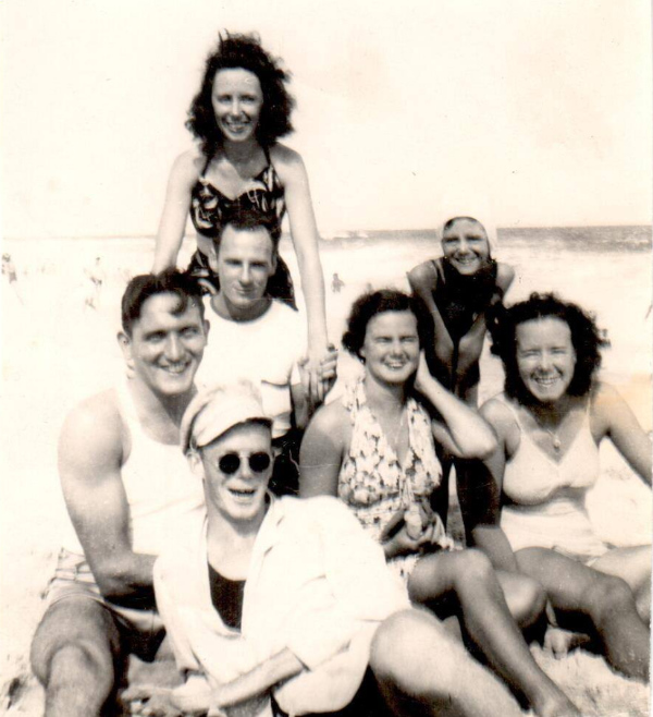 A midcentury black-and-white
photo with four women and three men, sitting on the beach and wearing midcentury beachwear. All are in their twenties
and thirties, it appears.