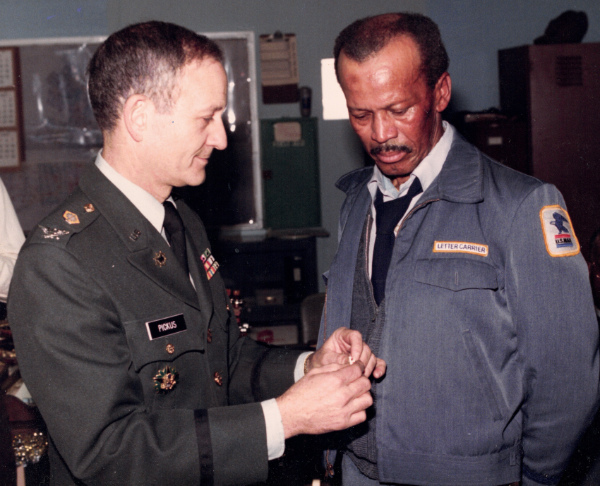 The picture
is as described. A middle-aged white fellow in a colonel's uniform delivers awards to a middle-aged Black fellow in a 
USPS uniform