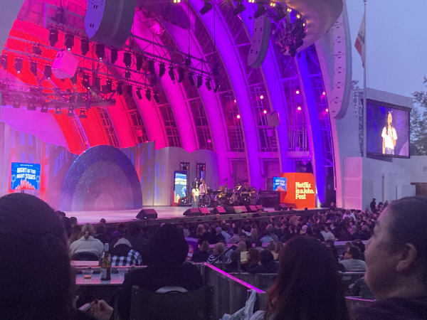 The Hollywood Bowl
is backlit in pink and magenta, Ali Wong is standing on the center of the stage, and she's also being projected 
on a giant TV next to the stage.