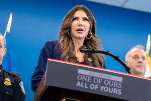 Kristi Noem stands at a podium;
the placard on front says 'ONE OF OURS ALL OF YOURS.'