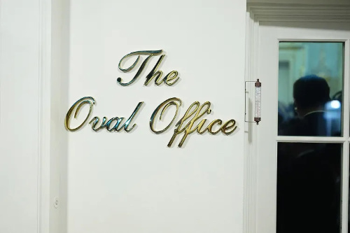 Ugly gold cursive 
letters spell out 'The Oval Office' on the wall outside the Oval Office