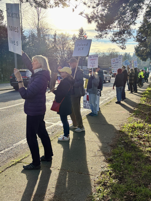 Several dozen people 
are visible, they are standing along the side of a road, and nearly all are holding up signs