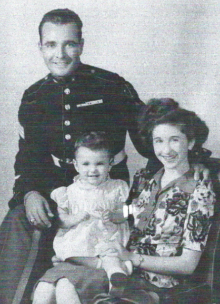 A black and white photograph
that looks to have been taken in 1946 or 1957. A handsome young woman, in period-fashionable clothing and hairstyle
is seated in an armchair, and is holding a young-ish child, who appears to be around 3 years old. Perched on the arm 
of the armchair is a dashing fellow in a formal Marine Corps uniform. The two adults look to be around 25 years of age.